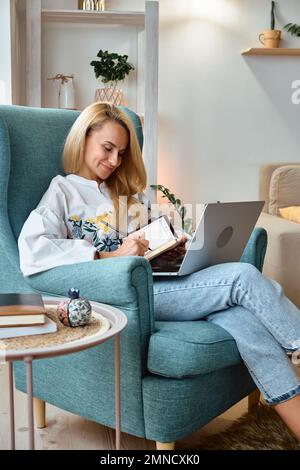 Giovane donna che usa il laptop e scrive in un notebook seduto in poltrona, lavoro a distanza a casa Foto Stock