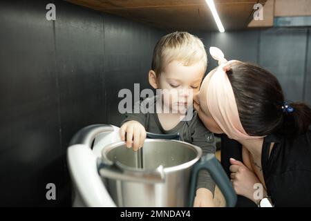 Mamma con amore bacia piccolo figlio sulla guancia. Il bambino guarda il vaso di metallo profondo del frullatore. Bambino piccolo aiutare madre cucinare cena. Foto Stock