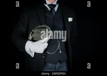 Ritratto di maggiordomo d'epoca in tuta scura e guanti bianchi che tiene elegantemente il vassoio di servizio in argento. Service Industry e cortesia professionale. Foto Stock