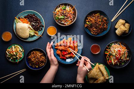 Set di piatti cinesi sul tavolo, le mani femminili che tengono bacchette: Pollo dolce e agrodolce, involtini primavera fritti, tagliatelle, riso, panini al vapore con barbecue Foto Stock