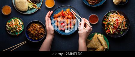 Set di piatti cinesi sul tavolo, le mani femminili che tengono bacchette: Pollo dolce e agrodolce, involtini primavera fritti, tagliatelle, riso, panini al vapore con barbecue Foto Stock