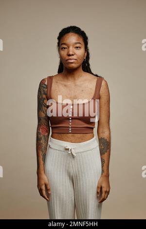 Ritratto minimo di giovane donna nera che guarda la macchina fotografica in piedi contro sfondo beige neutro con messa a fuoco sulla struttura della pelle reale con macchie e cicatrici acne Foto Stock