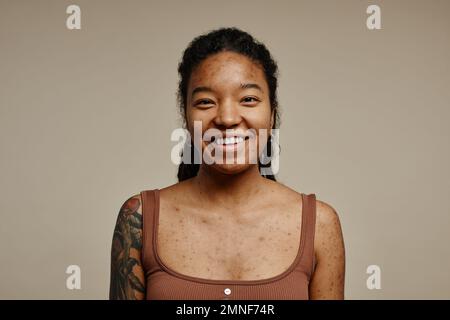 Ritratto minimo di giovane donna nera sorridente alla macchina fotografica in piedi contro lo sfondo beige neutro con messa a fuoco sulla struttura della pelle reale, macchie e cicatrici acne Foto Stock