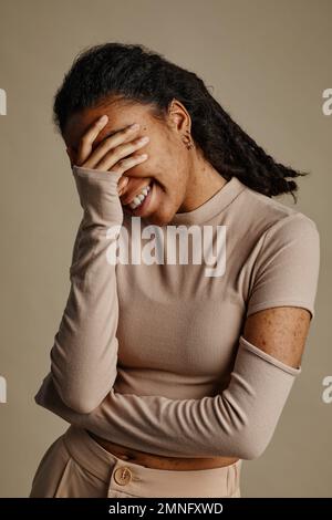 Ritratto verticale di giovane donna nera che ride sinceramente e coprendo il volto in tonalità neutre Foto Stock