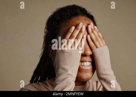 Primo piano di giovane donna nera sorridente felicemente e coprendo gli occhi con le mani, fuoco sulla struttura della pelle del viso con le cicatrici dell'acne, spazio della copia Foto Stock