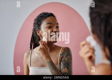 Ritratto minimale candida di giovane donna nera con cicatrici acne utilizzando il massaggiatore facciale durante la routine di bellezza e sorridente Foto Stock