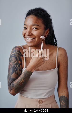 Ritratto verticale di giovane donna nera con cicatrici acne e tatuaggi utilizzando il massaggiatore facciale e sorridente, minimo Foto Stock