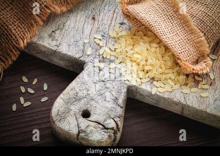 giallo riso marrone, usato per risotto in juta di sacco di burlap su un tagliere di legno Foto Stock