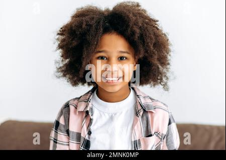 Ritratto di una bella ragazza afro-americana carina, età prescolare, con capelli ricci, in un vestito di base, siede sul divano, guarda alla macchina fotografica, sorrisi amichevole. Bambino felice Foto Stock