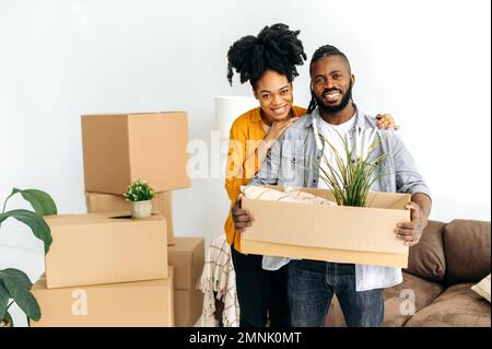 Gioiosi sposi afro-americani, trasferendosi in un nuovo appartamento, in piedi nel soggiorno, tenendo una scatola di cartone con le cose per la casa, guardando la macchina fotografica, sorridendo, felice insieme Foto Stock