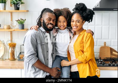 Amazing African American felice famiglia, papà mamma e la loro cute prescolare figlia, abbracciando insieme in una cucina a casa, guardare la macchina fotografica, sorridere, felice di trascorrere il tempo insieme, ragazza abbracciando i suoi genitori Foto Stock