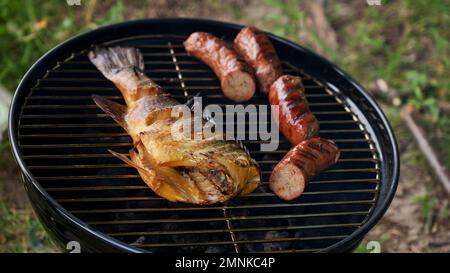 Vista dall'alto di pesce intero fresco alla griglia, spiedino, salsicce sulla griglia rotonda nera al carbone, sfondo verde erba. Barbecue, grill e concetto di cibo. Campin Foto Stock