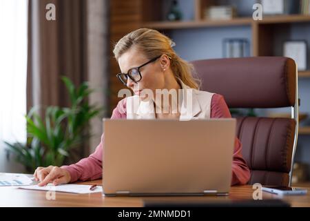 Ritratto di donna d'affari adulta con occhiali che lavorano in ufficio. Responsabile femminile che fa i documenti cartacei, analizza il rapporto finanziario, tiene i documenti Foto Stock
