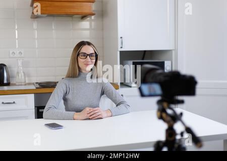 Blogger donna film video blog su videocamera professionale montato su treppiede. Allegra giovane adulta parla a casa con la fotocamera Foto Stock