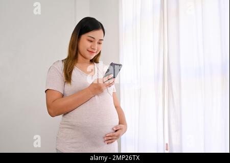 Felice donna asiatica incinta in abiti casual si trova accanto alla finestra utilizzando il suo smartphone e toccando il ventre. Foto Stock