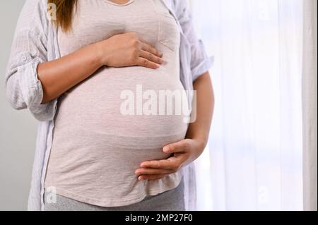 Una donna incinta in abiti comodi che tocca la pancia grande con amore e cura, sentendo il suo bambino nel pancino. immagine ritagliata Foto Stock