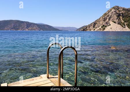 Scala in metallo al mare su molo in legno, vista pittoresca sulle montagne e sulla baia con acqua trasparente. Vacanza in spiaggia e concetto di nuoto Foto Stock