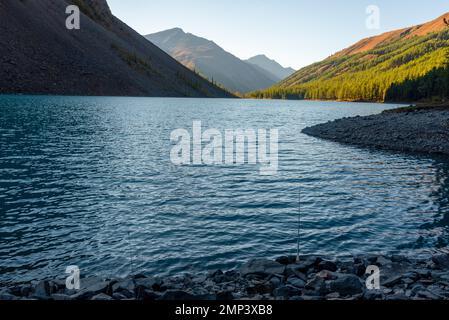 Una canna da pesca si trova all'ombra delle rocce sulla riva del lago alpino Shavlinskoe in serata. Foto Stock