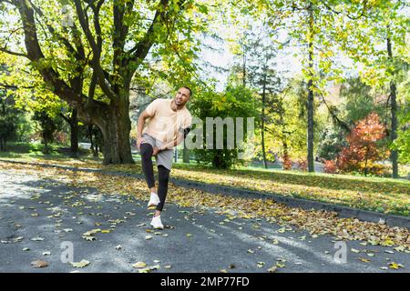 Giovane atleta afro-americano maschile che fa una corsa mattutina nel parco. Ferì il ginocchio, allungò i muscoli, si alza tenendo la gamba, sente dolore grave. Foto Stock