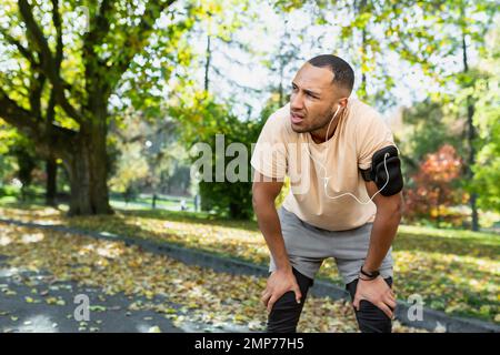 Un giovane atleta afro-americano ascolta la musica in cuffie bianche e va per una corsa mattutina nel parco. È stanco, fermato, respira duro, si alza e si riposa. Foto Stock