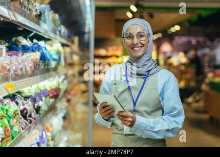 Una giovane donna araba in un hijab lavora in un supermercato, venditore, consulente, manager. In piedi con una cartella nelle mani e un badge nel reparto della drogheria. Guarda la telecamera, sorride. Foto Stock