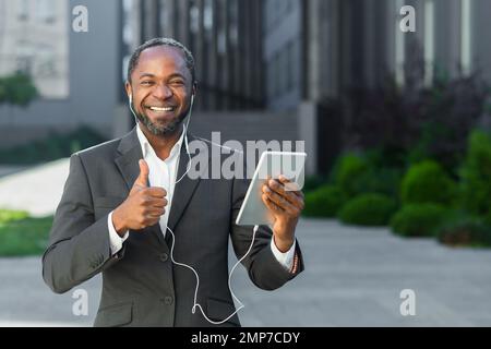 Un giovane afro-americano in tuta si trova all'esterno vicino a un centro ufficio che indossa le cuffie e tiene un tablet. Guarda nella fotocamera. sorrisi, punti con un super dito. Foto Stock