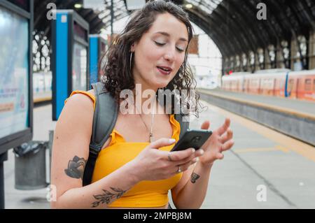 giovane donna latina viaggiatore al terminal dei treni che controlla il telefono felice perché finalmente hanno trasferito denaro, in piedi sul binario in attesa di Foto Stock