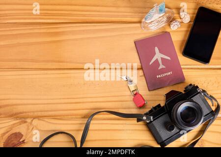 Vista dall'alto dell'attrezzatura essenziale da viaggio su un tavolo di legno. Passaporto e lucchetto chiusi su valigia giocattolo in miniatura, fotocamera e telefono cellulare. Assicurazione di viaggio Foto Stock