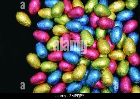 Mucchio di diversi colori e misure di foglio di cioccolato avvolto uova di pasqua in rosa, blu, giallo e verde calce su uno sfondo nero. Foto Stock