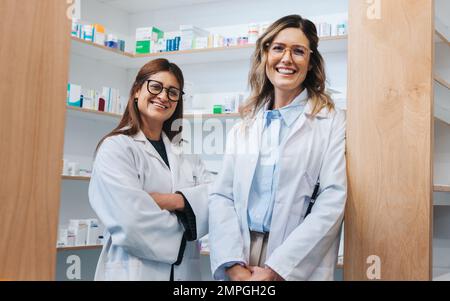 Farmacie femminili in piedi in una farmacia e sorridente alla telecamera. Due operatori sanitari che lavorano insieme in farmacia. Foto Stock