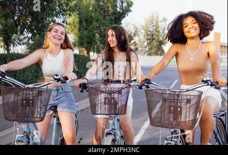 Adolescenti di diverse nazionalità e aspetto in bicicletta percorrono una strada cittadina. Le ragazze giovani e positive sorridono insieme. Stile di vita sano A. Foto Stock