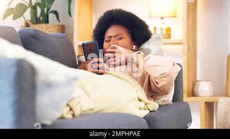 Donna stanca e annoiata sdraiata su un divano che digita sul telefono a casa. Una giovane ragazza africana che si rilassa e sbadigia su un divano in casa sua su un noioso Foto Stock