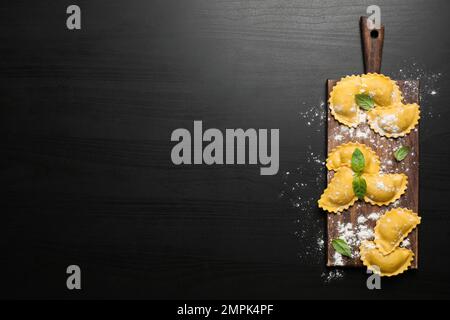 Ravioli crudi con basilico su tavola di legno nero, giacitura piatta e spazio per il testo. Pasta italiana Foto Stock