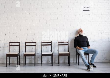 tutta la lunghezza dell'uomo maturo che dorme sulla sedia mentre aspetta il casting sulla sedia vicino alla parete bianca Foto Stock
