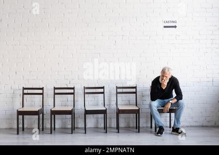 lunghezza intera di uomo pensivo in dolcevita nero e jeans in attesa di casting su sedia vicino parete bianca Foto Stock