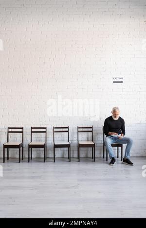 uomo di mezza età in dolcevita nero seduto su sedia e la lettura sceneggiatura mentre in attesa di casting Foto Stock