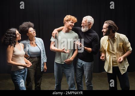 felice produttore che parla con l'uomo rosso vicino sorridente attori interrazziali sul palco del teatro Foto Stock