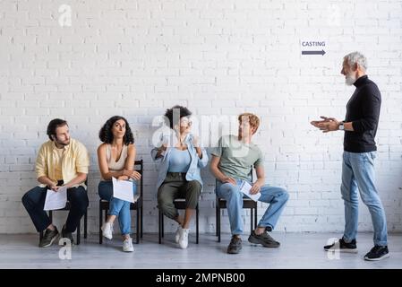 produttore dai capelli grigi che parla con felice afroamericana vicino a attori multietnici scettici in attesa di casting Foto Stock