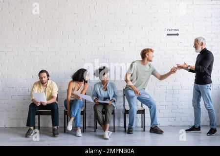 produttore bearded che indica l'attore di redhead vicino ai cercatori di lavoro interracial che aspettano per il getto nel corridoio Foto Stock