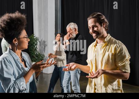 un uomo brunetta sorridente che parla con una donna afroamericana vicino a colleghi interrazziali nella scuola di recitazione Foto Stock