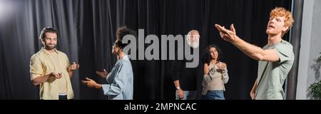 giovane uomo rosso in posa con mano tesa vicino attori interrazziali parlare in recitazione scuola, banner Foto Stock