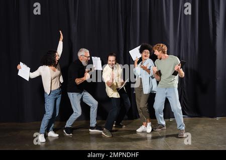 troupe teatrale multirazziale overjoyed con clipboard con sceneggiature e gesti di successo. Traduzione del tatuaggio: om, shanti, pace Foto Stock