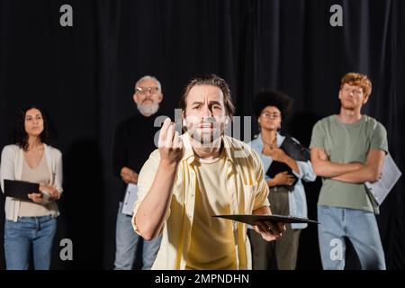 uomo emozionale con appunti che guarda la fotocamera e gesture durante le prove presso attori multietnici e produttore su sfondo sfocato Foto Stock