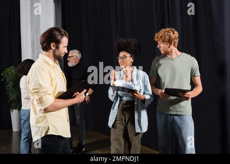 donna afroamericana sorridente con scenario che parla con i giovani attori nella scuola di abilità recitazione Foto Stock