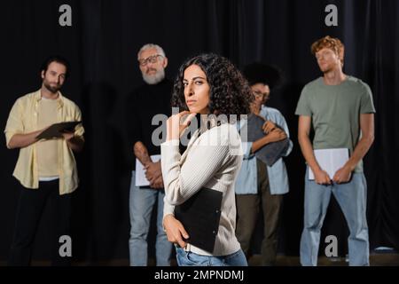 donna multirazziale con espressione del volto rigorosa che guarda la fotocamera mentre prova vicino al direttore d'arte e gli attori su sfondo sfocato Foto Stock