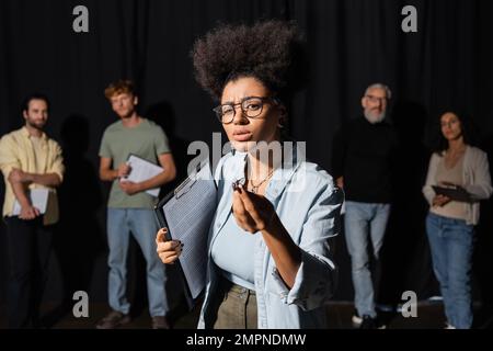 donna afroamericana che tiene la sceneggiatura e gesturing durante le prove vicino agli attori sfocati e all'insegnante di abilità comportanti Foto Stock