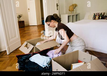 Vista laterale della donna che confezionava gli abiti in una scatola di cartone mentre si inginocchiava a casa Foto Stock