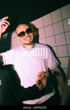 Giovane uomo sorridente che balla contro una parete bianca illuminata Foto Stock