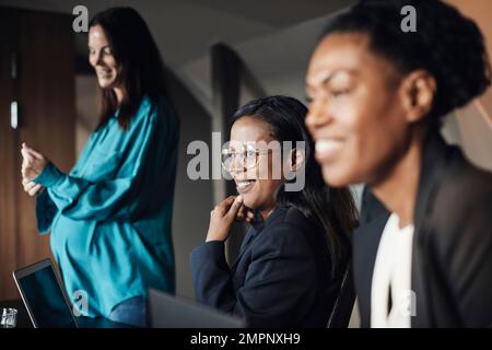 Donna d'affari sorridente con mano sul mento da parte dei colleghi durante la riunione in ufficio Foto Stock