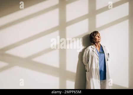 Donna medico indossare camice da laboratorio giorno sognando mentre si è in piedi contro la parete con luce solare Foto Stock